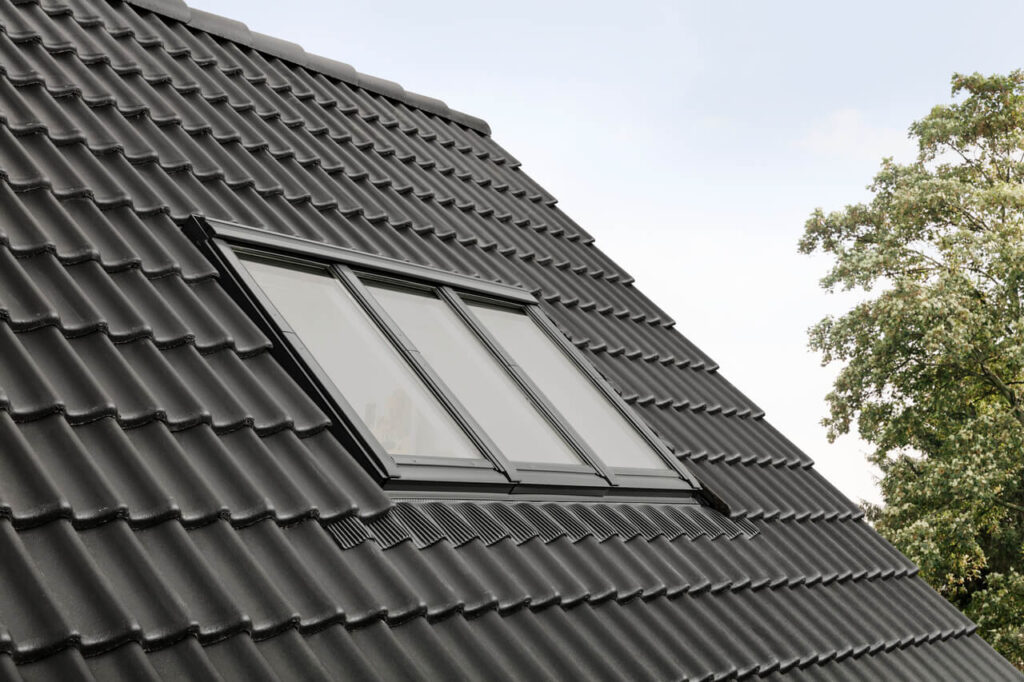 A close-up of a modern house featuring a Velux skylight window on a textured, dark tiled roof with a tree in the background.