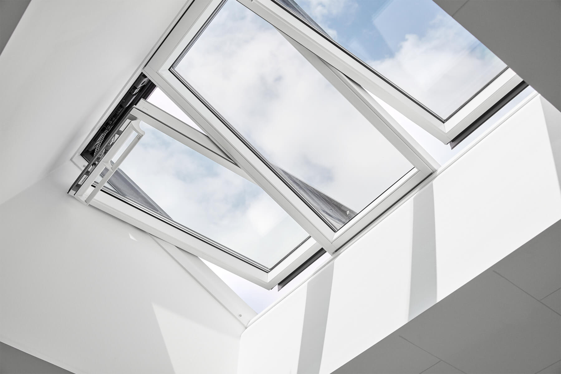 The image shows an open Velux skylight window set in a white ceiling with a view of a blue sky with clouds.