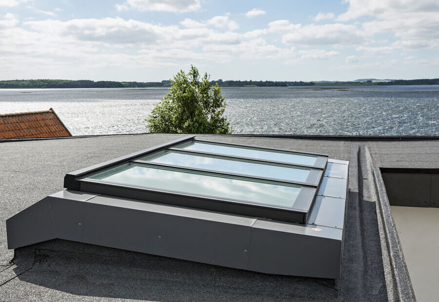 A flat roof window Velux skylight installed on a modern building with a view of a lake and sky in the background.
