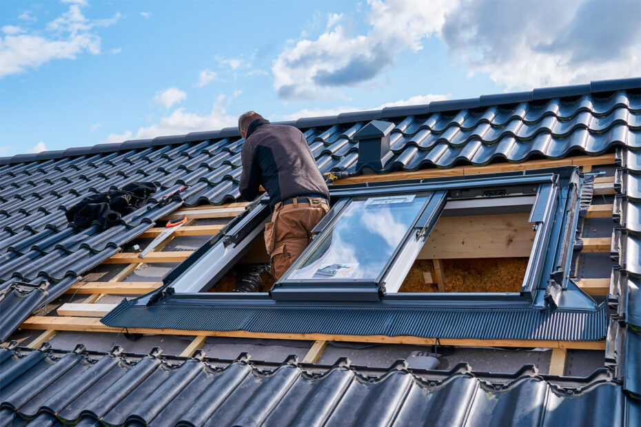 A worker is installing a Velux skylight on a tiled residential roof under a partly cloudy sky.