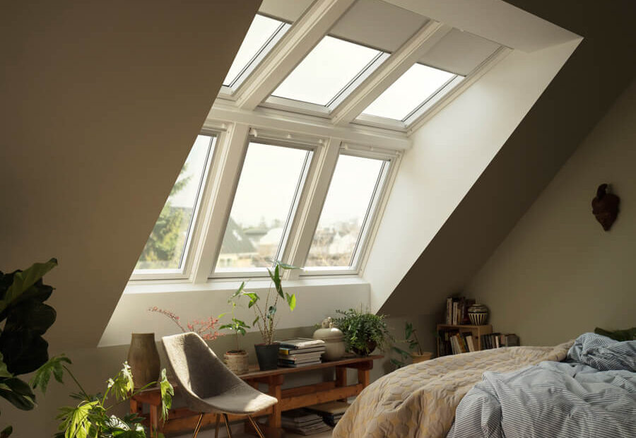 A cozy attic bedroom with Velux skylight windows, plants, a bed, and a reading nook, illuminated by natural daylight.