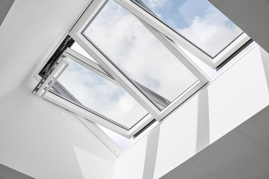 An open Velux skylight window in a minimalist white ceiling offers a view of a blue sky with fluffy clouds.