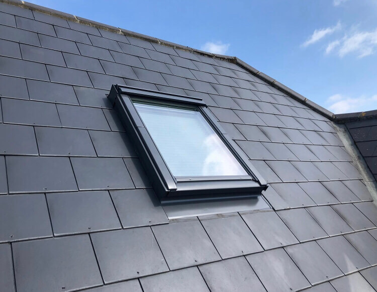 A Velux skylight window installed on a tiled sloping roof against a clear sky.