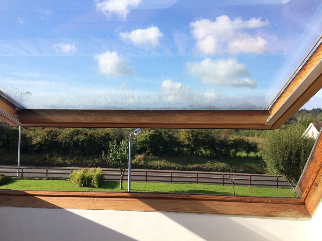 A Velux skylight window showing a view of blue sky with clouds, a green landscape, and a portion of a wooden balcony railing.