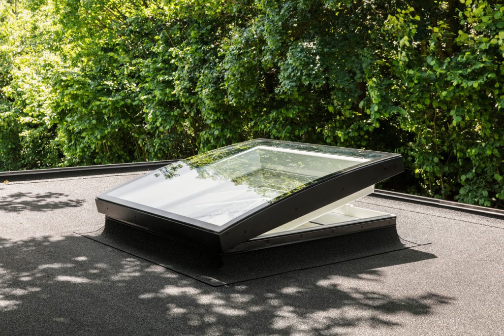 A Velux skylight window installed on a flat roof, surrounded by greenery reflecting on the glass.